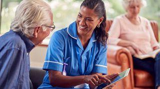 Healthcare practitioner holding a tablet is smiling whilst sitting with elderly man